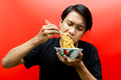 Portrait of Happy Asian man in black t-shirt eats instant noodles using chopsticks and bowl isolated on red background.