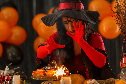 Copy space shot of an evil witch, a young woman in a face paint and costume, standing over a table with Halloween decorations, gesturing a spell, doing witchcraft and making sparks fly with the ignited sparkler.