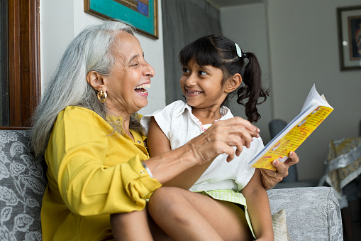 Old grandmother laughing while reading a book with her granddaughter at home