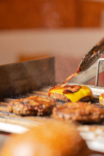 persona que agrega tocino a una hamburguesa con queso en una parrilla - hamburger burger symmetry cheeseburger fotografías e imágenes de stock