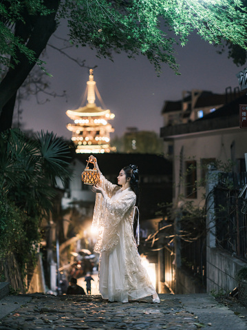 The girl in ancient Chinese costume is holding a lantern in front of the ancient pagoda in Hangzhou China.