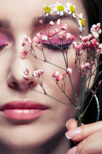 Closeup portrait of female face with pink makeup. Woman with Gypsophila or babe's breath flowers in hand near face. Brows are decorated by small chamomile flowers.  Eyes closed.