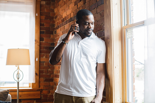 Portrait of smiling mid adult Black man standing by the window, looking outside and talking on the phone with a friend.