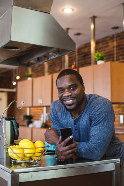 istock Charming Black man standing in his kitchen, leaning against the counter, using smart phone, smiling at camera 1423533187