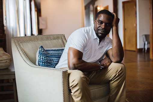 Three quarter length shot of smug mid adult Black man sitting in armchair, with head in hand, confidently looking at camera.