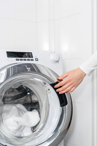closeup of female hand closing door in automatic washing machine with dirty clothes or bath towel in white bathroom. household appliance, housekeeping concepts
