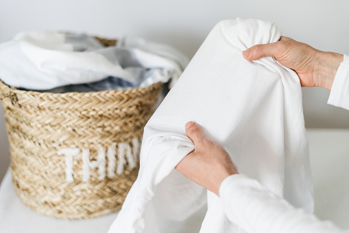selective focus on female hands folding fresh white clothes or dry cotton sheet after laundry near wicker basket in apartment room. concept of housework duties. woman checking stain