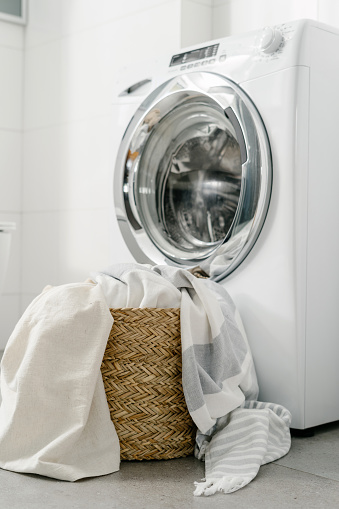full wicker basket with white linen bedspread and sheets on floor, close to empty automatic washing machine in modern bathroom in house. housework routine concept