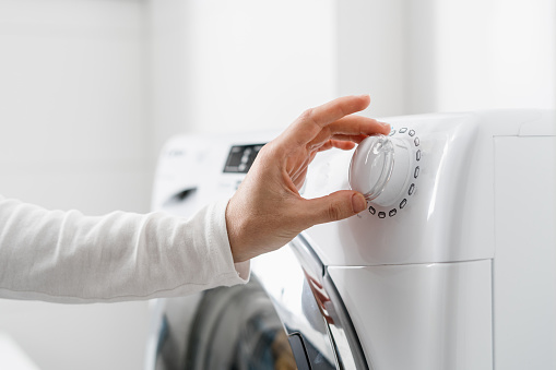 cropped shot of woman hand turn on automatic washing machine or select program with knob on control panel in white bathroom, modern appliances at home