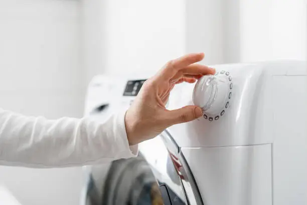 cropped shot of woman hand turn on automatic washing machine or select program with knob on control panel in white bathroom, modern appliances at home