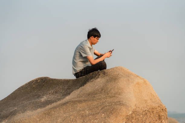 man sitting on a rock by the sea is using a smartphone