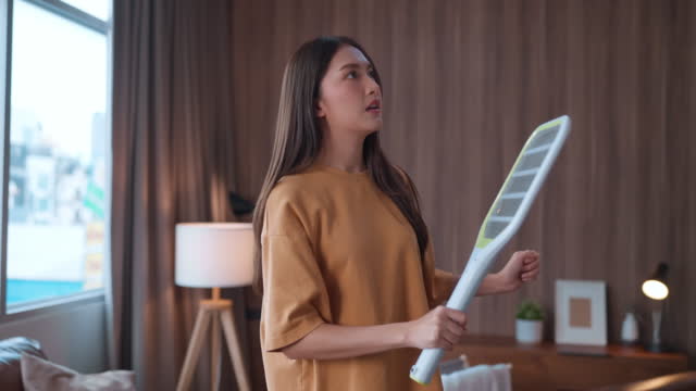 Young asian female woman using mosquito swatter to chasing annoy mosquito or fly bugs, electric net racket in living room near sitting sofa at home