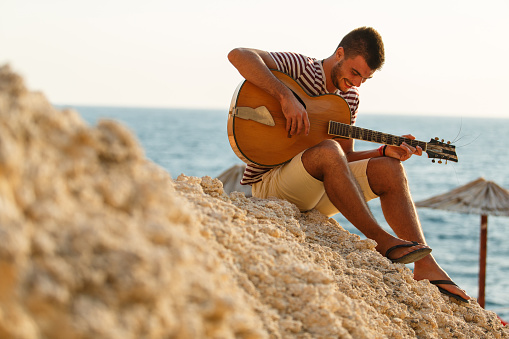Full length shot of joyful young man sitting on a rocky beach, enjoying summer sunshine and playing acoustic guitar.