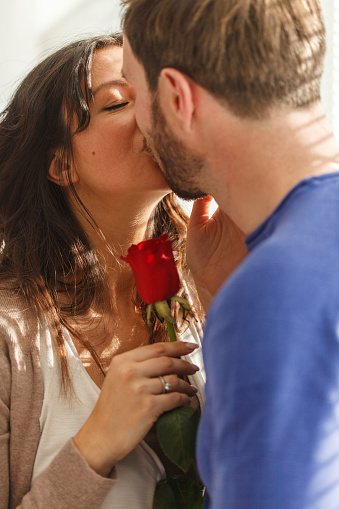 Close up shot of affectionate mid adult woman holding a single red rose and kissing her loving husband on the lips.