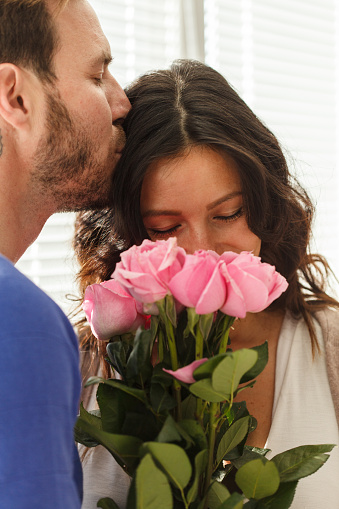 Close up shot of affectionate mid adult man kissing his wife while she is smelling a bouquet of roses he gave her for their anniversary.