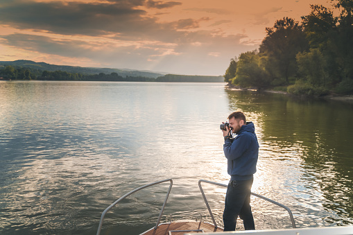 Young male photographer taking pictures of the sunset on a photo camera while traveling on a yacht on the river