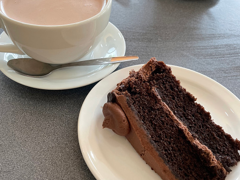 Stock photo showing close-up view of freshly made cup of hot chocolate, pictured on a table and ready to drink. The drinking chocolate is presented in a simple, stylish white china cup and saucer, and has been served alongside a slice of chocolate fudge cake that is displayed on a white plate.