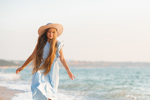 Beautiful smiling blonde teenager girl 12-14 year old with long hair wear hat and stylish elegant dress walk over sea coast outdoor. Summer season.