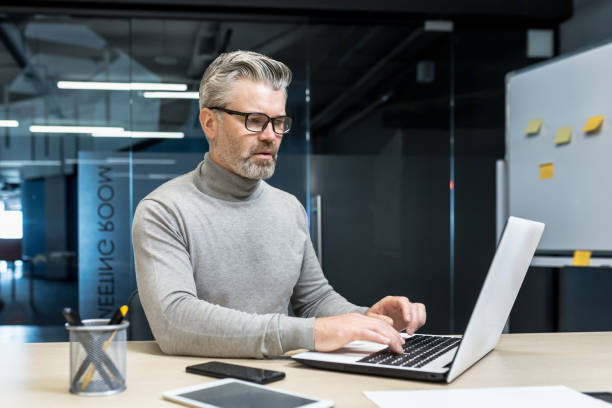 istock Senior and experienced programmer works in modern office at laptop, male web designer developer in glasses writes code programs new application, mature businessman works at desk 1423544106