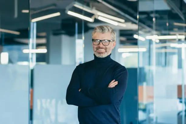 Portrait of happy gray-haired boss in glasses, businessman working in modern office standing by window smiling and looking at camera with crossed arms, satisfied investor in glasses