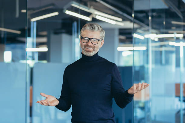 istock Senior gray-haired male businessman looking into web camera and smiling while standing in office, 1423544149
