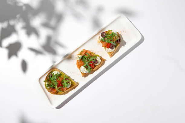 istock Aesthetic composition with salmon bruschetta on white background with shadows from flowers. Italian bruschetta with salmon, avocado, cheese and olives on fine dining in summer. Elegant menu concept. 1423548275