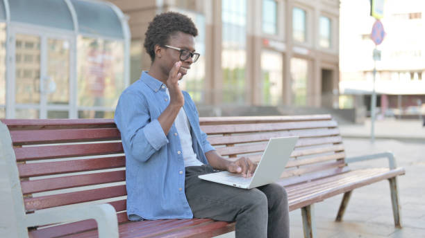 istock Young African Man Talking on Video Call while Sitting Outdoor on Bench 1423559496