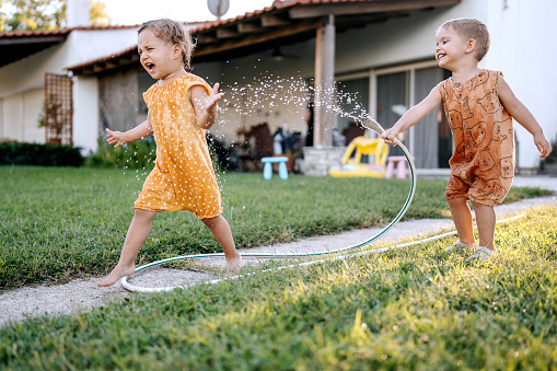 Two cute little siblings playing with a hose in a backyard, girl is crying and running away from her brother