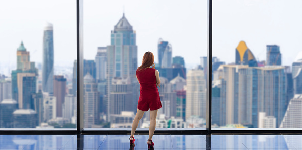 Luxury young Asian CEO woman entrepreneur looking at the downtown skyline at the window with skyscraper and cityscape for vision and real estate development concept