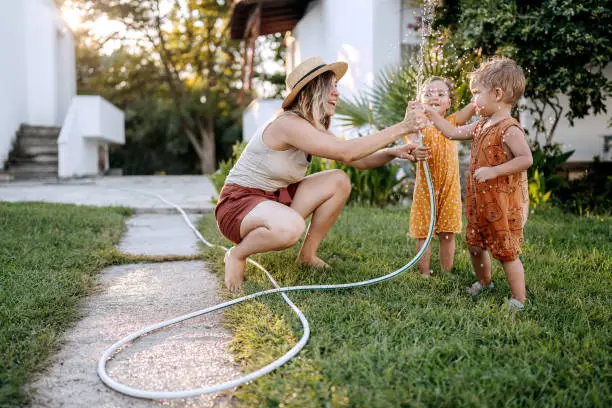 Mother and her cute little children playing with water with garden hose in a backyard