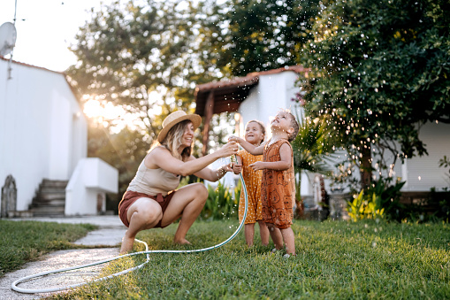 Mother and her cute little children playing with water with garden hose in a backyard