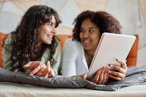 Digital tablet in hands of young African American woman talking to her girlfriend while both relaxing on comfortable double bed