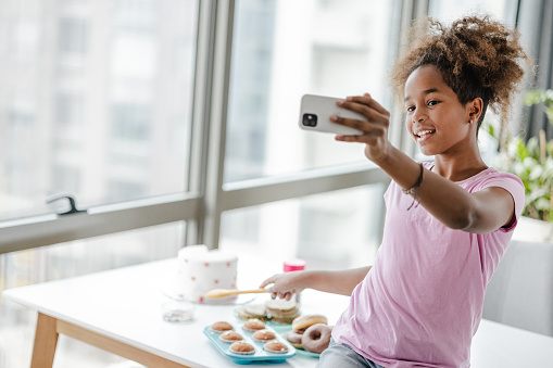 African American teenage girl having fun in the kitchen while vlogging. She is sitting on the table and using smart phone.