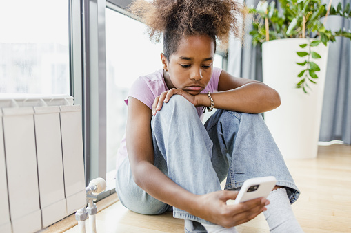 African-American girl is at home by the window. She is holding a mobile phone