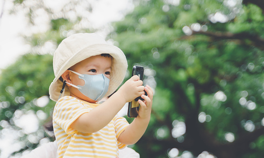 Smiling Small Asian Toddler boy wearing hat and protective face mask taking selfie on smartphone at nature outdoor, Little Child using mobile phone in a park