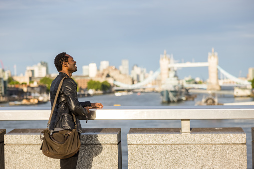 Cheerful mature Black male walking in London, looking at the Tower Bridge and the Thames River. He is happy, laughing and smiling.