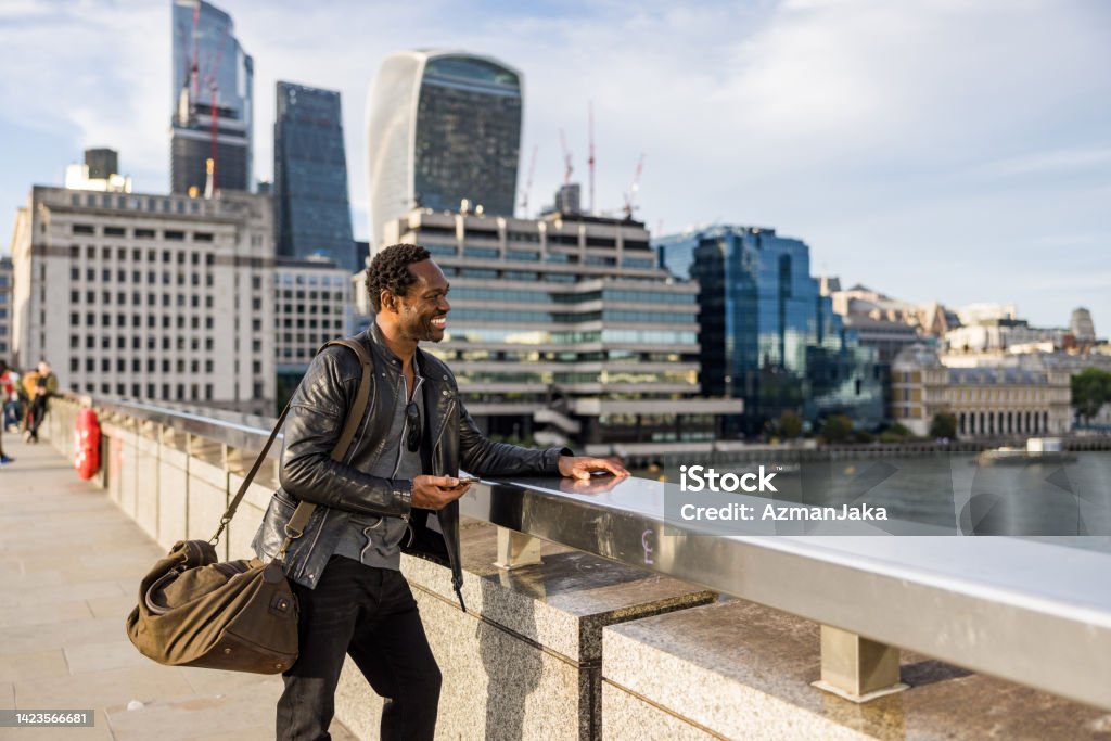 Cool Mature African American Male Traveler Looking For Adventure In London Happy African American male solo traveler enjoying a cityscape view from a bridge across a river. He is wearing casual clothing and carrying a big bag. Commercial buildings in the background. 50-54 Years Stock Photo
