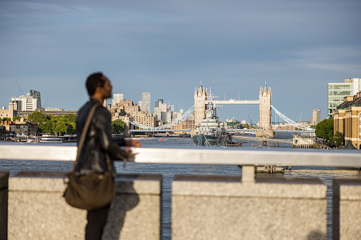 Young African American male tourist appreciating the view of the Tower Bridge in London on a sunny day. He is standing and wearing casual clothing.