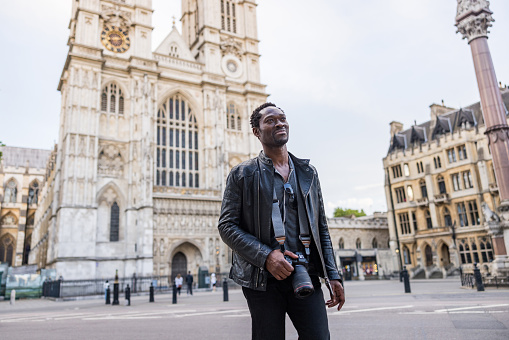 Young African American male traveling and discovering London and it's beautiful architecture. He is holding a professional camera while walking in front of the Westminster Abbey facade.