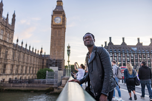 Handsome mature African American male tourist standing on a bridge across River Thames. He is visiting the Big Ben and other famous places in Central London. He is looking away.