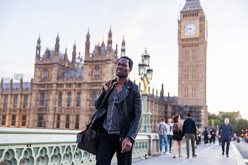 Black male exploring London city on foot. He is wearing casual clothes and carrying a bag. He is looking away, walking on a bridge in front of the Big Ben tower.