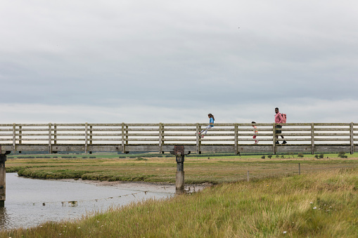 A wide angle side view of a father and his two children crossing a bridge whilst on a staycation in Beadnell in the North East of England. The kids are swinging along holding on to the sides and being safely supervised by their father.
