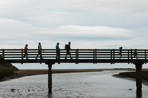 A side view silhouette of a family on a staycation to the beach in Beadnell in the North East of England. The adults are walking behind and the two children are running ahead over the bridge towards the beach.