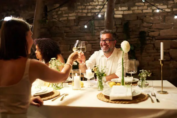 Cheerful mature man and young female guest clinking with wineglasses over served festive table while enjoying wedding party