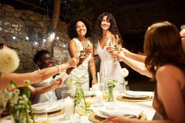 Two happy young intercultural brides toasting with their wedding guests Two happy young intercultural brides toasting with their wedding guests and clinking with flutes of champagne over served festive table wedding feast stock pictures, royalty-free photos & images