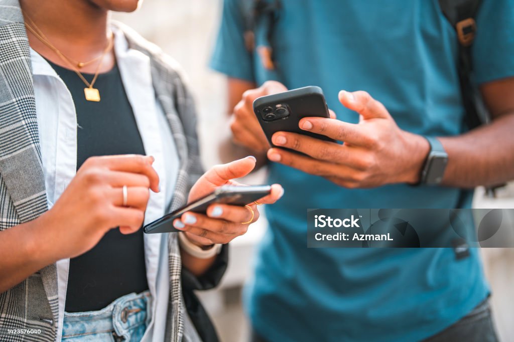 Handsome Young Black Couple Using Smart Phones While Hanging Out In The City Beautiful young African American couple walking in the city, scrolling on their smart phones and hanging out. They are wearing casual clothing. Using Phone Stock Photo