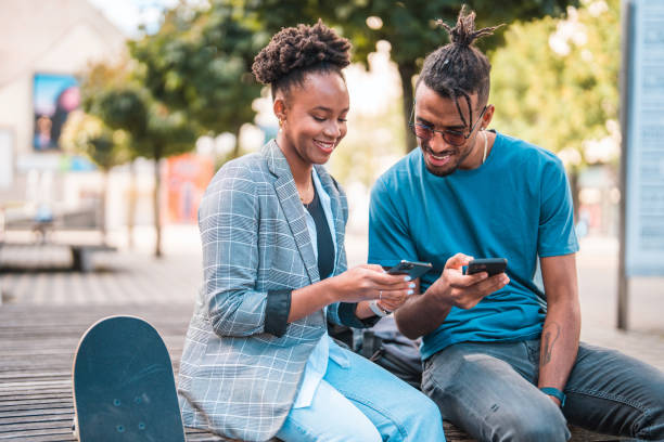 casal jovem e negro bonito sentado em uma bech ao ar livre, usando seus telefones inteligentes - bech - fotografias e filmes do acervo