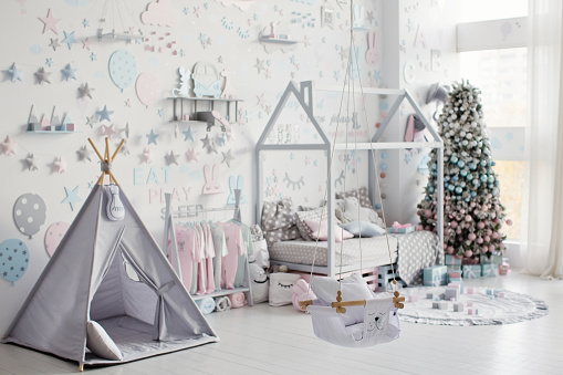 Cute kids room interior with house shaped bed, grey wigwam, blue and pink decorations on the white wall at Christmas time. Beautiful Christmas tree in childs room