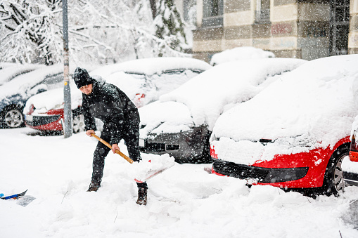 A Man with Snow Shovel is Shoveling a Snow in Front of Car During a Cold Snowy Winter Day.