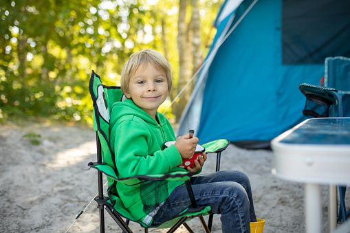 Cute child, having breakfast in front of pitched tent on the beach, while wild camping in Norway, summertime
